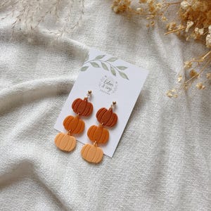 May include: A pair of pumpkin-shaped earrings in shades of orange and brown. The earrings are attached to a white card with the text "Eden & Ivy Designs" and are displayed on a textured, off-white surface.