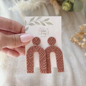 May include: Terracotta-colored, patterned arch earrings with a matching top stud. The earrings are held against a white card with the text "Eden & Ivy Designs" and a floral design. The earrings have a geometric design.