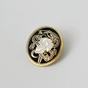 May include: A black and gold enamel pin featuring a skull with a snake wrapped around it. The snake is biting its own tail, forming a circle. The skull is white with black details. The pin is circular and has a gold border.