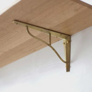 May include: A gold-colored metal shelf bracket is mounted on a light-colored wooden shelf. The bracket has a decorative curved support and is attached to the shelf with screws. The wall is white.