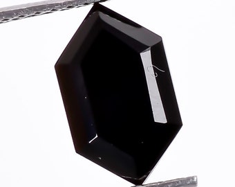 May include: A faceted, black gemstone held by silver tweezers against a white background. The gemstone has a hexagonal shape with polished surfaces that reflect light. The gem is a deep, opaque black color.