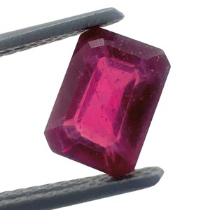May include: A faceted, rectangular ruby gemstone held by grey tweezers. The ruby has a deep, saturated pink colour and is set against a white background. The gemstone's cut and clarity are visible.