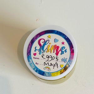 Pode incluir: Um recipiente branco com um r&oacute;tulo colorido que diz "Slime" e tem um arco-&iacute;ris, estrelas e cora&ccedil;&otilde;es. O r&oacute;tulo tamb&eacute;m diz "Nome: eggs" e "Por: maya".