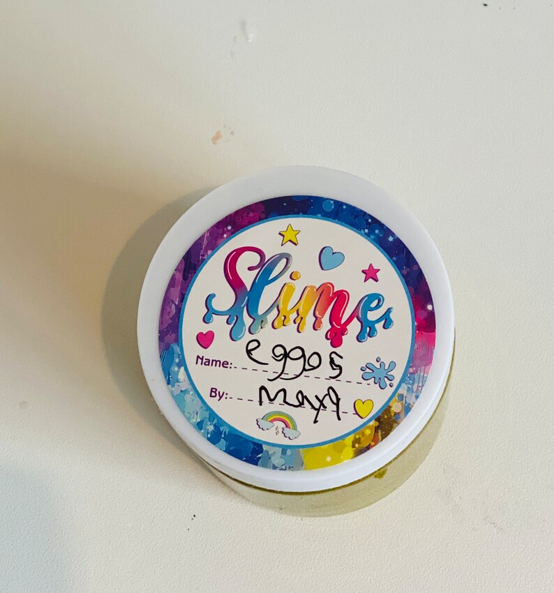 Pode incluir: Um recipiente branco com um r&oacute;tulo colorido que diz "Slime" e tem um arco-&iacute;ris, estrelas e cora&ccedil;&otilde;es. O r&oacute;tulo tamb&eacute;m diz "Nome: eggs" e "Por: maya".
