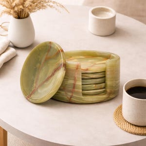 May include: A set of green onyx coasters with a matching holder. The coasters have a natural, veined pattern with streaks of red and brown. A cup of coffee sits on a woven coaster next to the set.