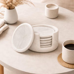 May include: A set of white marble coasters with a matching holder. The coasters are oval-shaped and stacked neatly in a round holder. A single coaster rests on the side. A cup of coffee and a small vase are also on the table.
