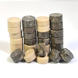 Può includere: Una collezione di pedine da gioco in pietra nei toni del beige e del grigio. I pezzi sono cilindrici con una parte superiore e inferiore leggermente concave. Alcuni pezzi sono impilati in colonne, altri sparsi.