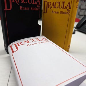 Puede incluir: Tres ejemplares del libro Dr&aacute;cula de Bram Stoker. Uno est&aacute; en un estuche negro con texto rojo. Un segundo ejemplar tiene una cubierta amarilla con texto rojo. Un tercero es blanco con texto rojo y un borde rojo en la portada.