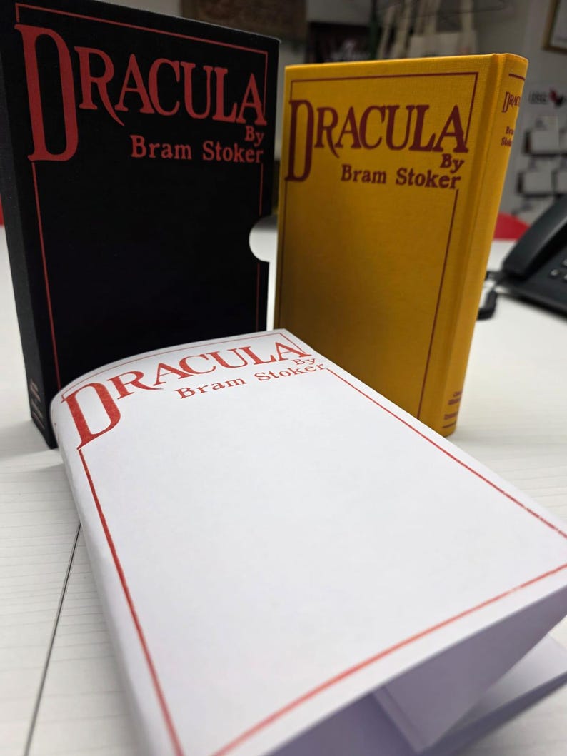 Puede incluir: Tres ejemplares del libro Dr&aacute;cula de Bram Stoker. Uno est&aacute; en un estuche negro con texto rojo. Un segundo ejemplar tiene una cubierta amarilla con texto rojo. Un tercero es blanco con texto rojo y un borde rojo en la portada.