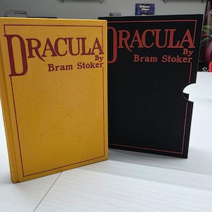 Puede incluir: Un libro de tapa dura amarillo titulado Dracula de Bram Stoker junto a un estuche negro con el mismo t&iacute;tulo en letras rojas. El libro amarillo tiene un borde y texto rojos, mientras que el estuche negro tiene un contorno y texto rojos.