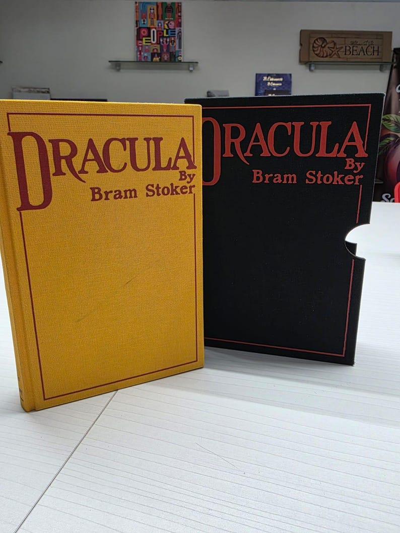 Puede incluir: Un libro de tapa dura amarillo titulado Dracula de Bram Stoker junto a un estuche negro con el mismo t&iacute;tulo en letras rojas. El libro amarillo tiene un borde y texto rojos, mientras que el estuche negro tiene un contorno y texto rojos.