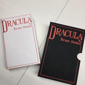 Puede incluir: Dos ejemplares del libro Dr&aacute;cula de Bram Stoker. Uno tiene una cubierta blanca con texto y borde rojos. El otro es un estuche negro con texto y borde rojos, que presenta un recorte semicircular en el lateral.