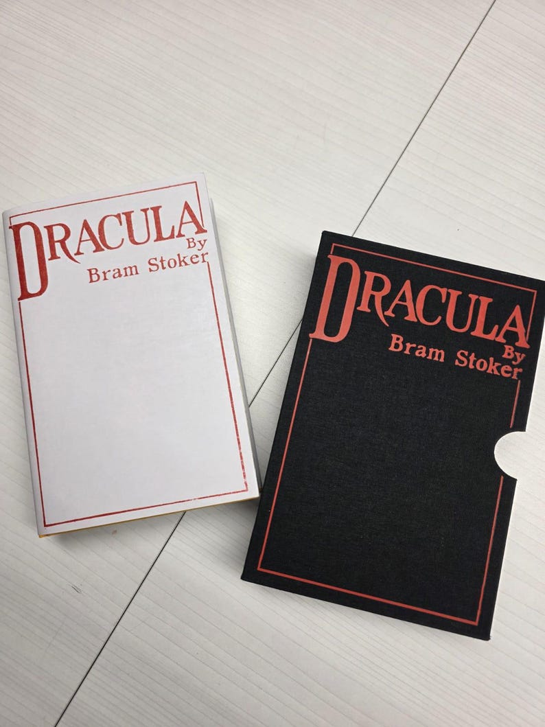 Puede incluir: Dos ejemplares del libro Dr&aacute;cula de Bram Stoker. Uno tiene una cubierta blanca con texto y borde rojos. El otro es un estuche negro con texto y borde rojos, que presenta un recorte semicircular en el lateral.