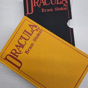 Puede incluir: Un libro de tapa dura color amarillo mostaza titulado Dracula de Bram Stoker, con texto y borde en rojo. Detr&aacute;s se encuentra un estuche negro con el mismo t&iacute;tulo y nombre del autor impresos en rojo, incluyendo una muesca.