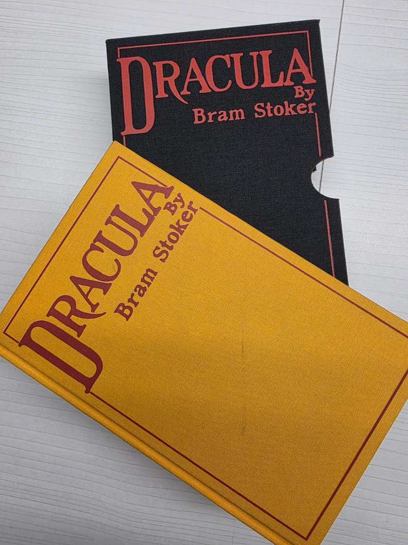 Puede incluir: Un libro de tapa dura color amarillo mostaza titulado Dracula de Bram Stoker, con texto y borde en rojo. Detr&aacute;s se encuentra un estuche negro con el mismo t&iacute;tulo y nombre del autor impresos en rojo, incluyendo una muesca.