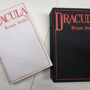 Puede incluir: Un libro blanco titulado Dracula de Bram Stoker con texto y borde rojo, junto a un estuche negro que presenta el mismo t&iacute;tulo y autor en texto rojo con un borde rojo y una muesca lateral para facilitar la extracci&oacute;n del libro.