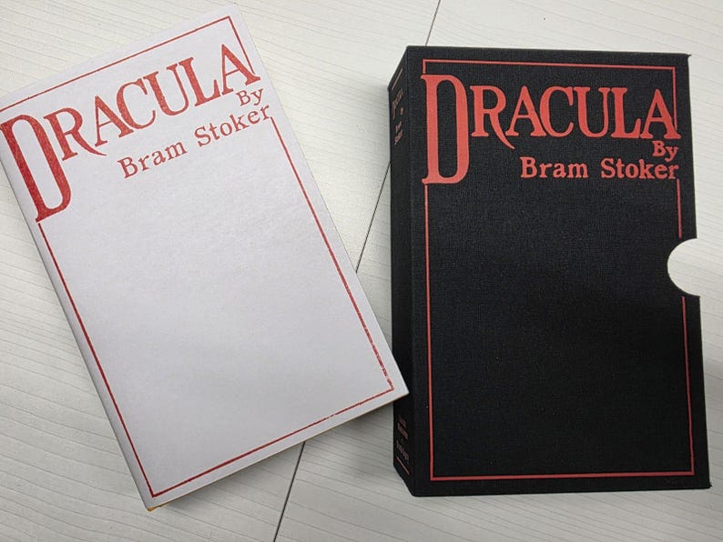 Puede incluir: Un libro blanco titulado Dracula de Bram Stoker con texto y borde rojo, junto a un estuche negro que presenta el mismo t&iacute;tulo y autor en texto rojo con un borde rojo y una muesca lateral para facilitar la extracci&oacute;n del libro.