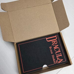 Puede incluir: Un libro negro titulado Dracula de Bram Stoker con letras y borde rojos, colocado dentro de una caja de env&iacute;o de cart&oacute;n marr&oacute;n abierta. La cubierta del libro presenta un recorte semicircular en el borde inferior para facilitar su extracci&oacute;n.