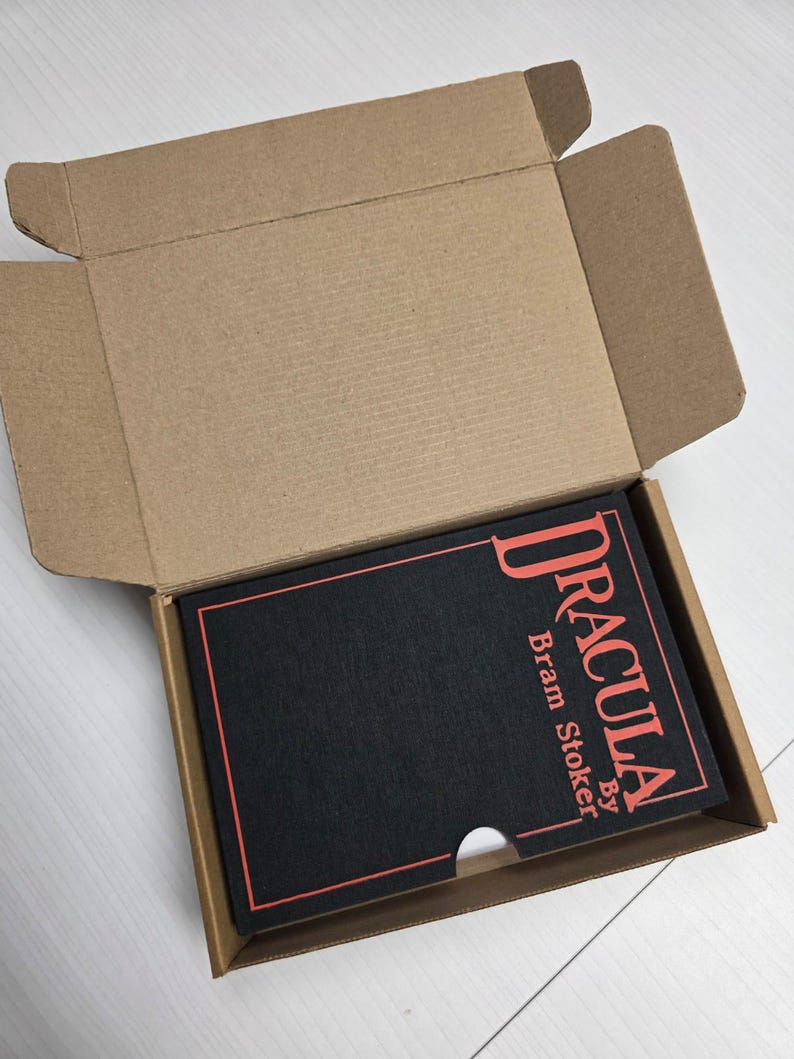 Puede incluir: Un libro negro titulado Dracula de Bram Stoker con letras y borde rojos, colocado dentro de una caja de env&iacute;o de cart&oacute;n marr&oacute;n abierta. La cubierta del libro presenta un recorte semicircular en el borde inferior para facilitar su extracci&oacute;n.
