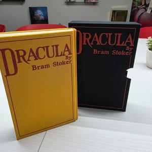 Puede incluir: Un libro de tapa dura amarillo titulado Dracula de Bram Stoker con letras y borde rojos, junto a un estuche negro a juego que presenta el mismo t&iacute;tulo y autor en texto rojo con un borde de contorno rojo.