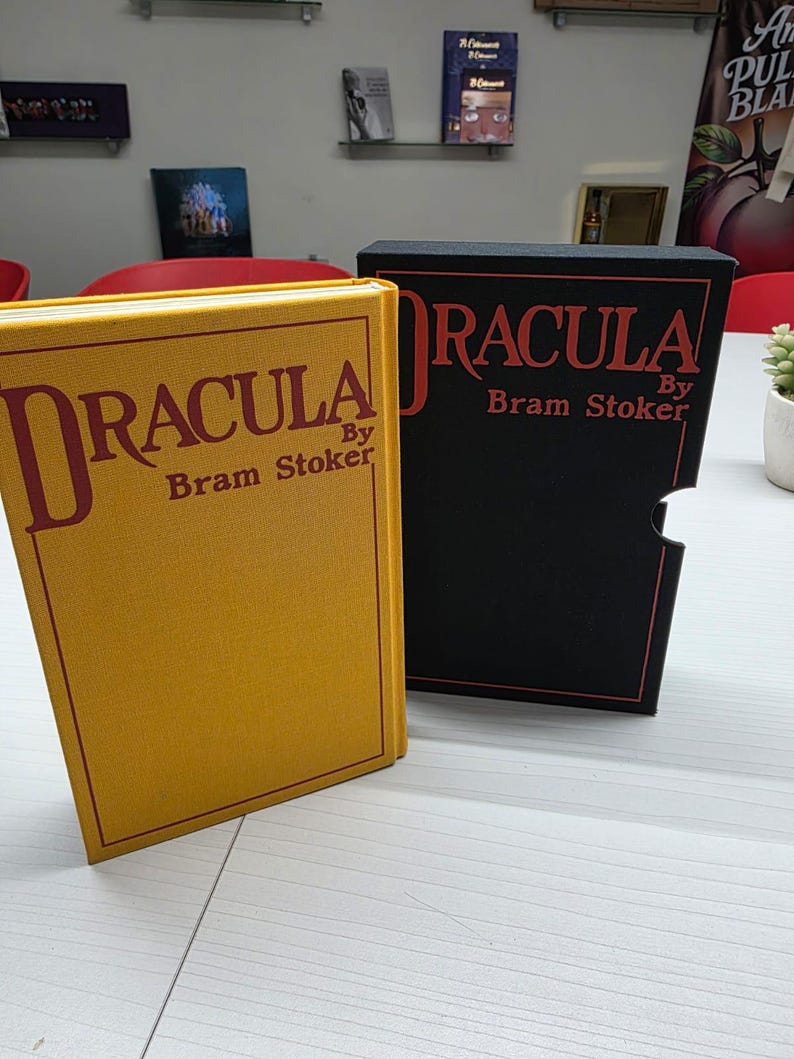 Puede incluir: Un libro de tapa dura amarillo titulado Dracula de Bram Stoker con letras y borde rojos, junto a un estuche negro a juego que presenta el mismo t&iacute;tulo y autor en texto rojo con un borde de contorno rojo.