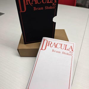 Puede incluir: Un estuche negro y una cubierta de libro blanca, ambos con el texto rojo Dracula By Bram Stoker dentro de un fino borde rectangular rojo. El estuche negro tiene un recorte semicircular en el lado derecho y una caja de cart&oacute;n marr&oacute;n detr&aacute;s.