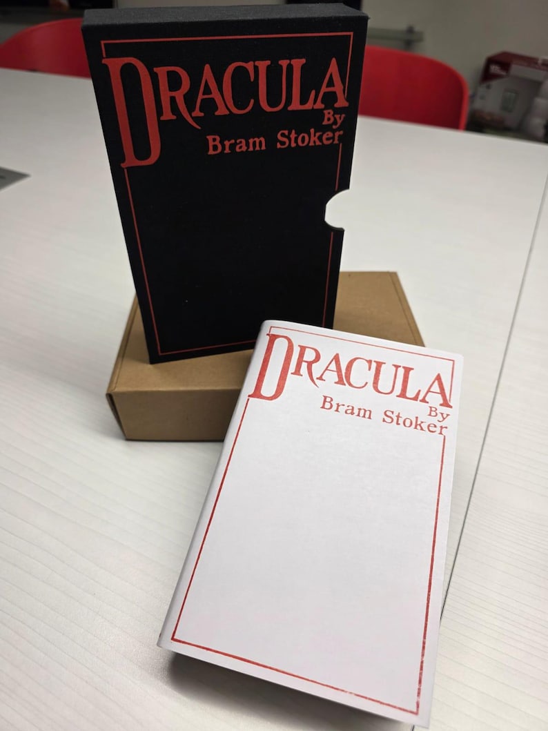 Puede incluir: Un estuche negro y una cubierta de libro blanca, ambos con el texto rojo Dracula By Bram Stoker dentro de un fino borde rectangular rojo. El estuche negro tiene un recorte semicircular en el lado derecho y una caja de cart&oacute;n marr&oacute;n detr&aacute;s.