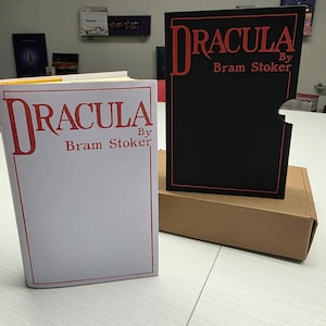 Puede incluir: Un libro blanco titulado Dracula by Bram Stoker con letras y borde rojos, junto a un estuche negro con el mismo t&iacute;tulo y autor en rojo, sobre una caja de cart&oacute;n marr&oacute;n colocada en una superficie blanca.