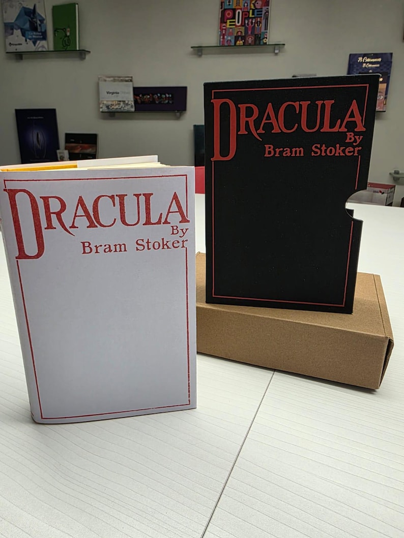 Puede incluir: Un libro blanco titulado Dracula by Bram Stoker con letras y borde rojos, junto a un estuche negro con el mismo t&iacute;tulo y autor en rojo, sobre una caja de cart&oacute;n marr&oacute;n colocada en una superficie blanca.