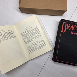 Puede incluir: Un ejemplar de tapa dura de Dr&aacute;cula de Bram Stoker con un estuche negro que presenta texto en rojo, junto a un libro abierto que muestra p&aacute;ginas de texto impreso y una caja de cart&oacute;n marr&oacute;n simple en la parte superior.