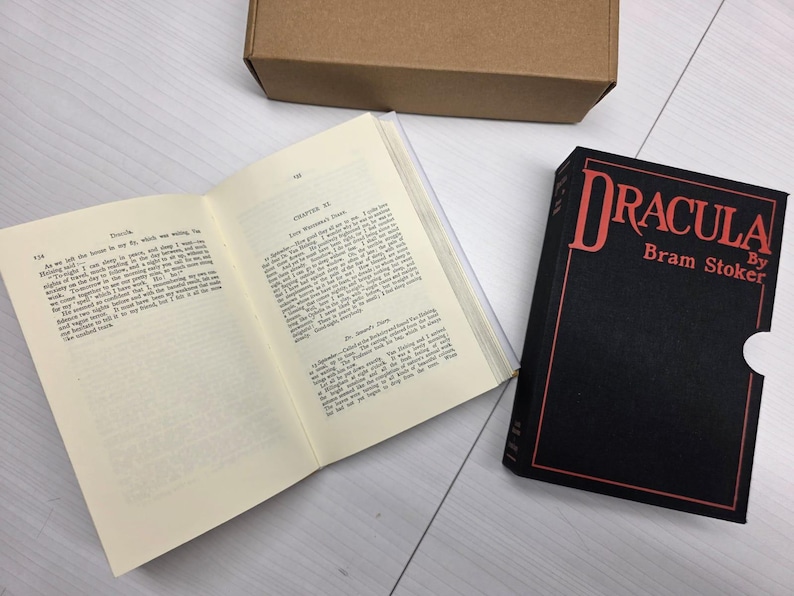 Puede incluir: Un ejemplar de tapa dura de Dr&aacute;cula de Bram Stoker con un estuche negro que presenta texto en rojo, junto a un libro abierto que muestra p&aacute;ginas de texto impreso y una caja de cart&oacute;n marr&oacute;n simple en la parte superior.