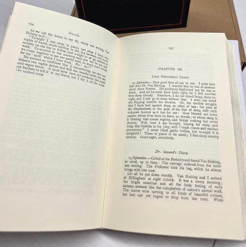 Puede incluir: Un libro de tapa dura abierto que muestra las p&aacute;ginas 134 y 135 de la novela Dr&aacute;cula. El texto incluye el cap&iacute;tulo XI, el diario de Lucy Westenra y el diario del Dr. Seward, impresos en tinta negra sobre papel blanco con tipograf&iacute;a serif.