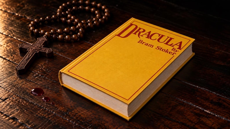 Puede incluir: Un libro de tapa dura amarillo titulado Dracula de Bram Stoker con letras y borde rojos. Junto al libro hay un crucifijo de madera marr&oacute;n oscuro con una cadena de rosario de cuentas y tres peque&ntilde;as manchas rojas circulares.