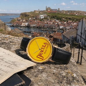 May include: A wristwatch with a black leather strap and a round gold-tone case. The watch face is yellow with a red border and features the text 'DRACULA by Bram Stoker' in a dark red font, along with three thin gold-tone hands for timekeeping.
