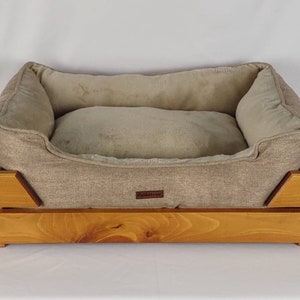 May include: A beige dog bed with a wooden frame. The bed has a removable cushion and a brown label that says "Cuddletown".