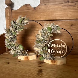Könnte beinhalten: Zwei Holzkränze mit getrockneten Blumen und Grün, einer mit einem schwarzen Metallring und einem Holzschild mit der Aufschrift "Anni & Florian" mit einem Herzen.