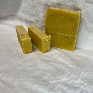 May include: Three bars of handmade soap in varying sizes. The soap has a light yellow-green color and a slightly textured surface. The bars are arranged on a white, textured cloth. The largest bar is rectangular, while the smaller bars are cut.