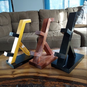 May include: Three wooden phone stands in different colors: yellow, brown, and black. Each stand has a unique angled design and a flat base.