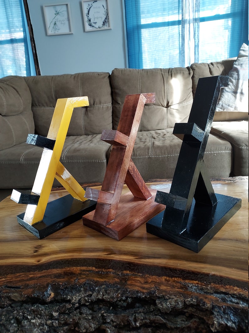 May include: Three wooden phone stands in different colours: yellow, brown, and black. Each stand has a unique angled design and a flat base.
