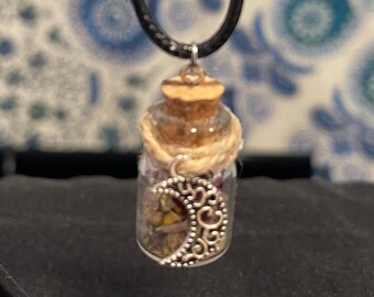 May include: A small glass bottle pendant necklace filled with dried herbs and flowers. The bottle has a cork stopper and is wrapped with twine. A silver crescent moon charm hangs below the bottle. The necklace cord is black.