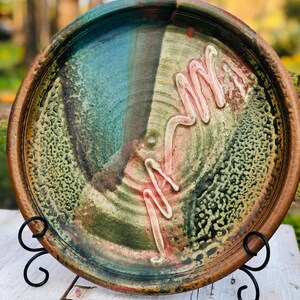May include: A handcrafted ceramic plate with a unique, abstract design. The plate features a blend of colors, including blue, green, and brown, with a textured surface and a raised, wavy white design. The plate is displayed on a black metal stand.