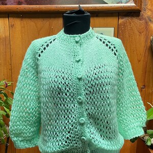 May include: A light green, hand-knitted cardigan with a round neckline and button closure. The sleeves are three-quarter length and the knit pattern features openwork detailing. The cardigan is displayed on a black mannequin.