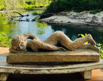 May include: A wooden sculpture of a reclining figure, with a rough, textured surface. The sculpture is placed on a wooden base, and the background shows a river with trees on either side.