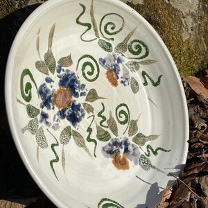 May include: White ceramic bowl with a floral design in shades of green, blue, and brown. The bowl has a slightly raised rim and a smooth, glazed surface.