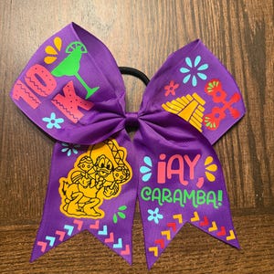 Può includere: Fiocco per cheerleader viola con elastico nero. Il fiocco presenta grafica e testo colorati, tra cui un margarita, una piramide e la frase "¡Ay, Caramba!". Il fiocco ha un design di personaggi dei cartoni animati.