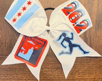 May include: White hair bow featuring the Chicago flag, the number 26.2, a runner silhouette, and the word "Chicago". The bow is made of a ribbon material and is attached to a black elastic band.