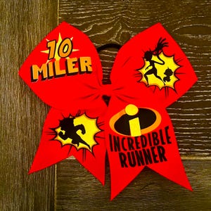 Puede incluir: Lazo de animadora rojo con un diseño negro y amarillo. El lazo presenta el texto "10 Miler" y "Incredible Runner" con una silueta de una figura corriendo.
