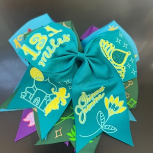Pode incluir: Um laço de cabelo verde-azulado com texto em glitter amarelo que diz 13.1 miles e Princess Jasmine. O design inclui uma silhueta de palácio em glitter amarelo, uma flor amarela e um gráfico de coroa, impressos em uma base de fita.