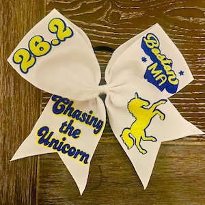 May include: White cheer bow with blue and yellow glitter lettering. The bow says "26.2", "Chasing the Unicorn", and "Boston MA". A yellow glitter unicorn is on one side of the bow.
