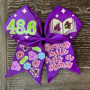Puede incluir: Lazo de animadora morado con un número 48,6 de purpurina verde, un libro morado y blanco con un castillo y un dragón verde, y el texto "Every mile has a story" en purpurina rosa. El lazo está decorado con gemas brillantes de colores.